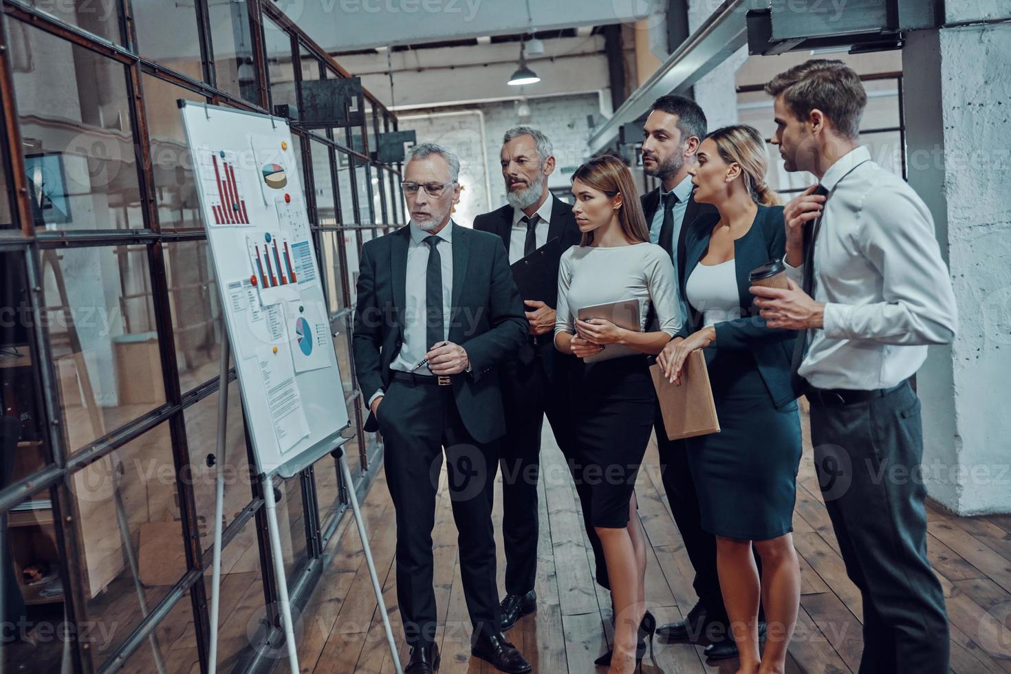 moderner geschäftsmann, der diagramm auf dem flipchart zeigt, während er eine mitarbeiterversammlung im bürokorridor abhält foto