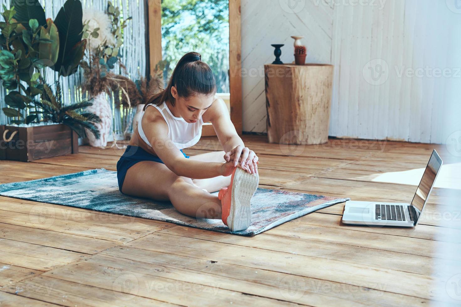 Schöne junge Frau in Sportkleidung, die sich auf der Terrasse mit Laptop in ihrer Nähe ausdehnt foto