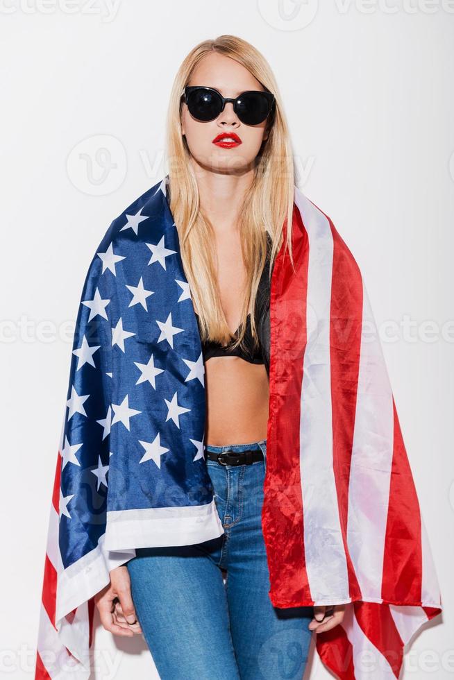 Amerikanischer Stil. Attraktive junge Frau in schwarzem BH und Lederjacke, die ihre Schultern mit amerikanischer Flagge bedeckt, während sie vor weißem Hintergrund steht foto