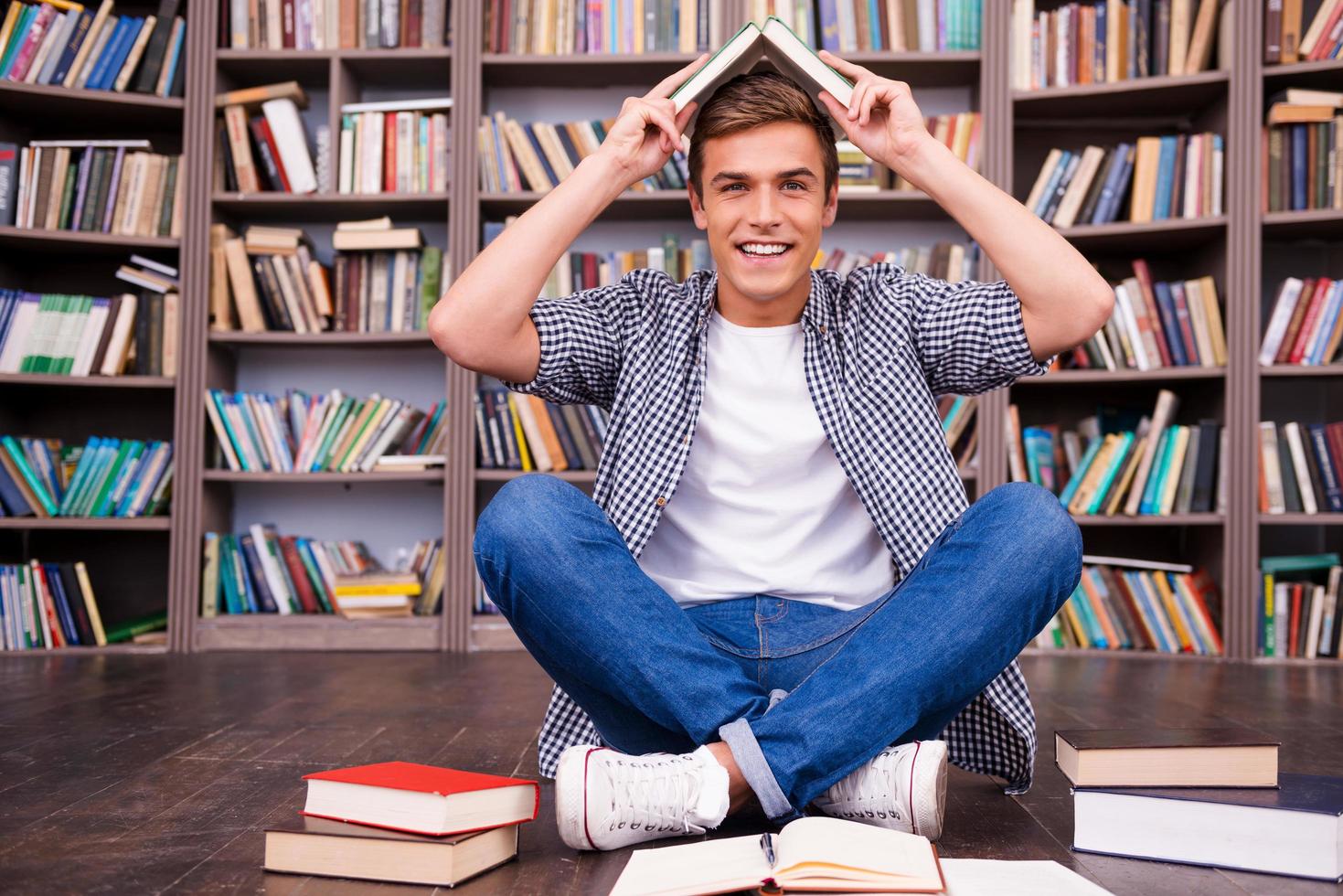 verspielter Schüler. Verspielter junger Mann, der ein Buch auf dem Kopf trägt und lächelt, während er gegen ein Bücherregal sitzt foto