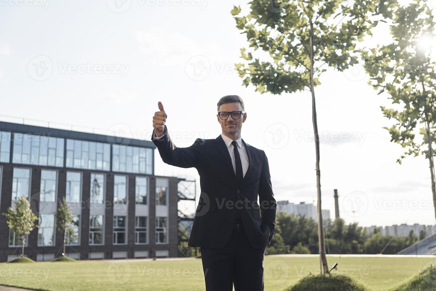 Selbstbewusster, reifer Geschäftsmann mit Brille, der seinen Daumen nach oben zeigt, während er im Freien steht foto
