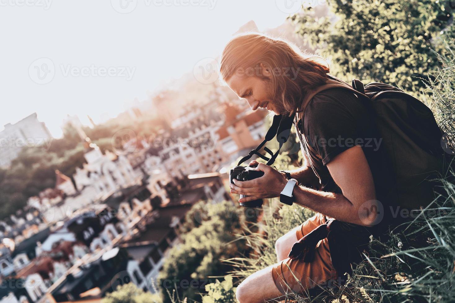 das Leben ist voller Schönheit. Junger Mann in Freizeitkleidung mit Kamera und lächelnd, während er draußen auf dem Hügel sitzt foto