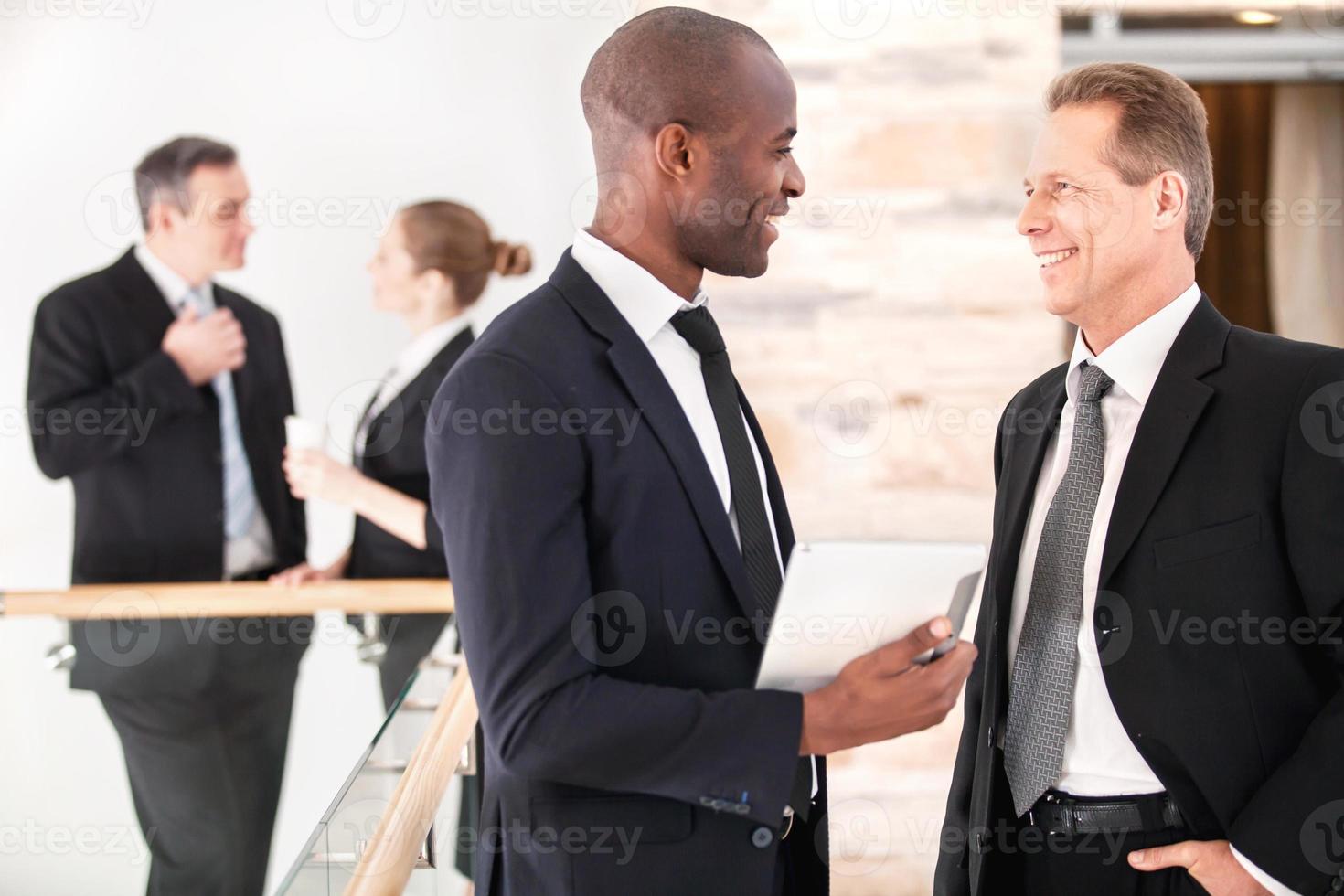 Unternehmenskommunikation. Zwei fröhliche Geschäftsleute reden miteinander, während ihre Kollegen im Hintergrund stehen foto
