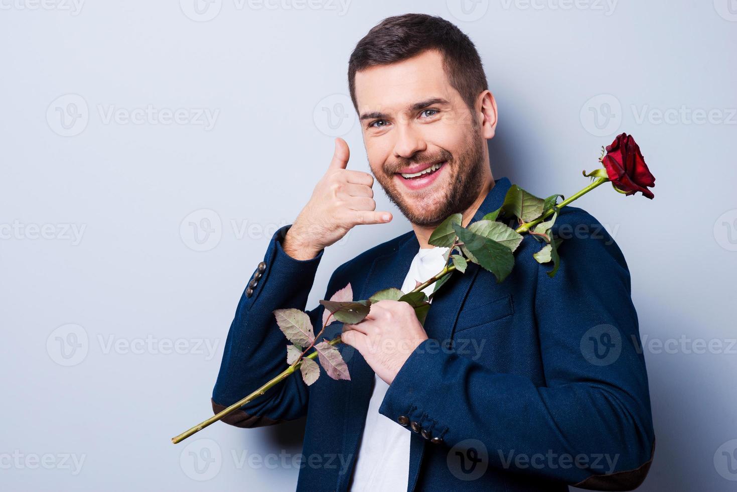 ich werde dich anrufen. hübscher junger mann, der rote rose auf schultern trägt und handy nahe gesicht gestikuliert, während er vor grauem hintergrund steht foto