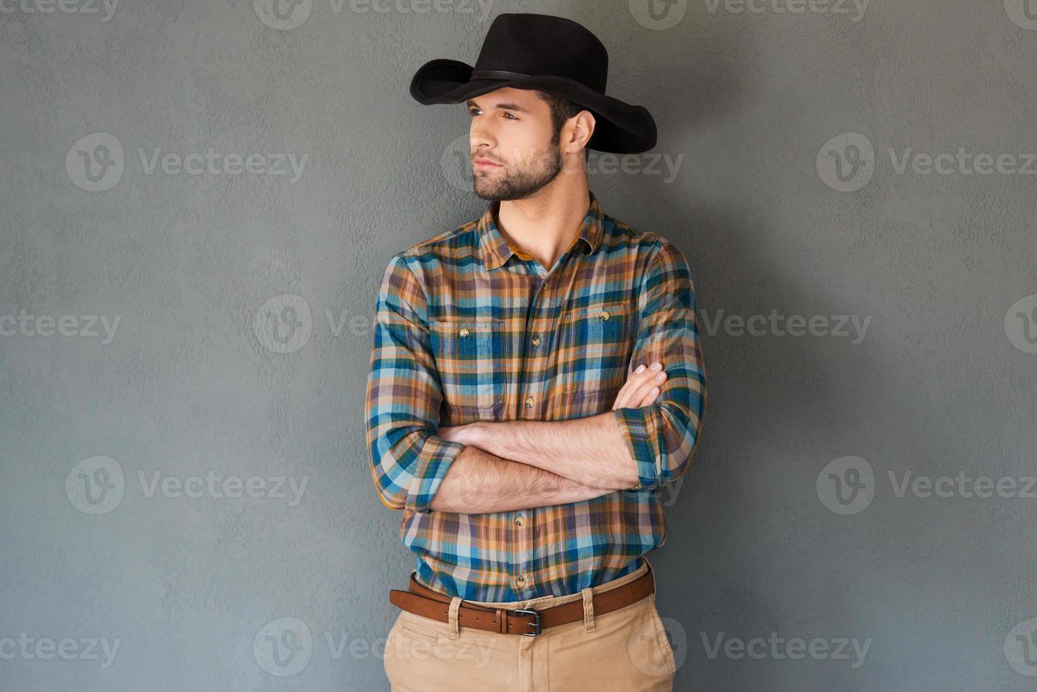 ernsthafter und selbstbewusster Cowboy. hübscher junger Mann mit Cowboyhut, der die Arme verschränkt hält und wegschaut, während er vor grauem Hintergrund steht foto