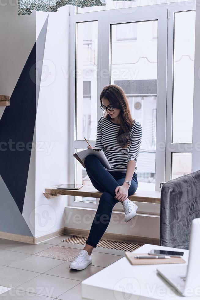 Ideen aufschreiben. nachdenkliche junge frau, die etwas in ihr notizbuch schreibt, während sie auf der fensterbank sitzt foto