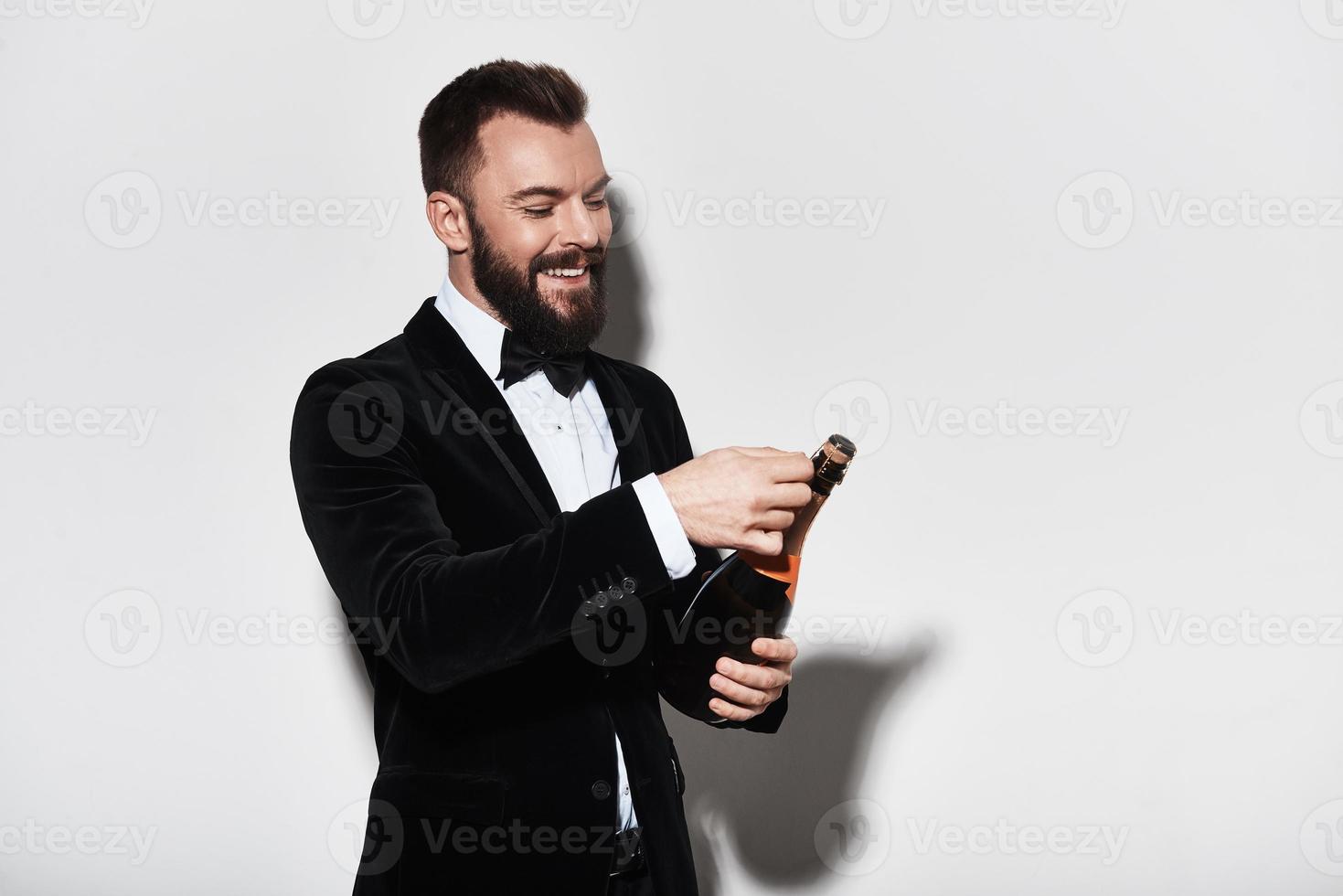 Lassen Sie uns einen hübschen jungen Mann im vollen Anzug feiern, der eine Flasche Champagner öffnet und lächelt, während er vor grauem Hintergrund steht foto