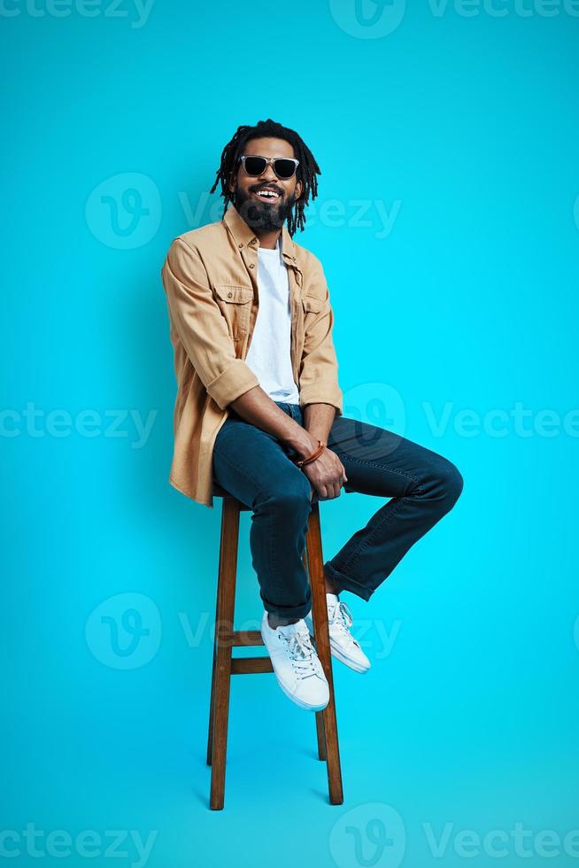 in voller Länge von einem hübschen jungen afrikanischen Mann in Freizeitkleidung, der in die Kamera schaut und lächelt, während er auf einem Hocker vor blauem Hintergrund sitzt foto