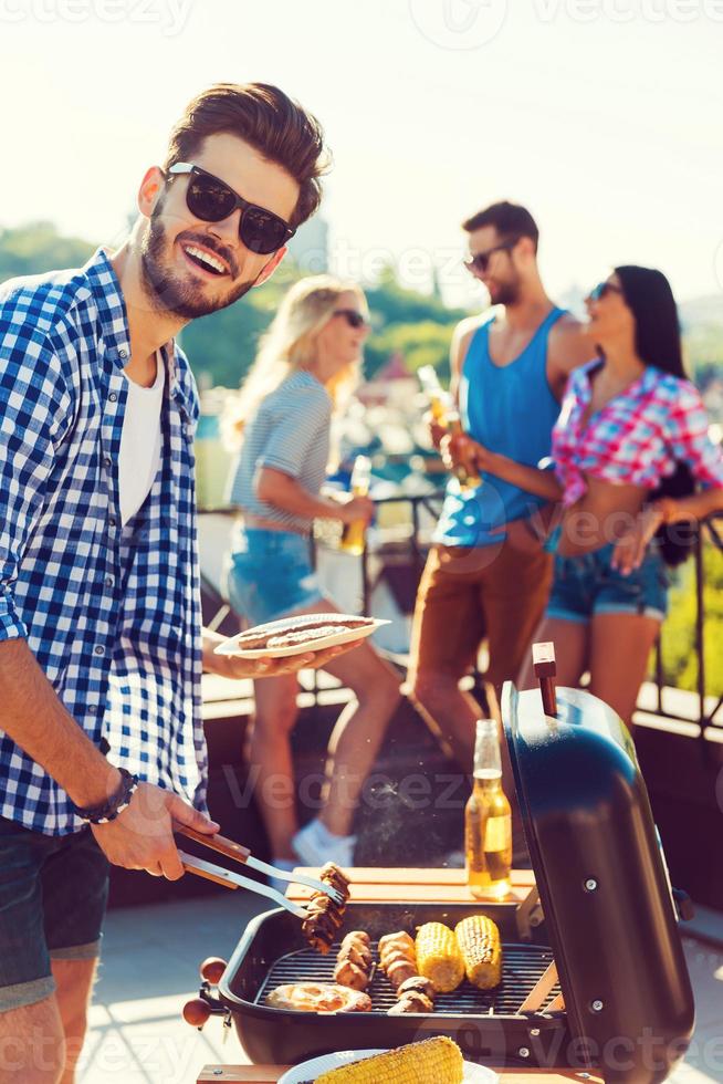 leckeres Essen und gute Gesellschaft. glücklicher junger Mann grillt und schaut in die Kamera, während drei Leute Spaß im Hintergrund haben foto