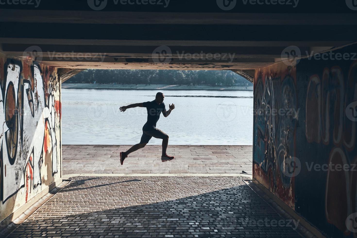 Weitsprung. Volle Länge des jungen afrikanischen Mannes in Sportkleidung, der beim Training in der Nähe des Flusses im Freien joggt foto