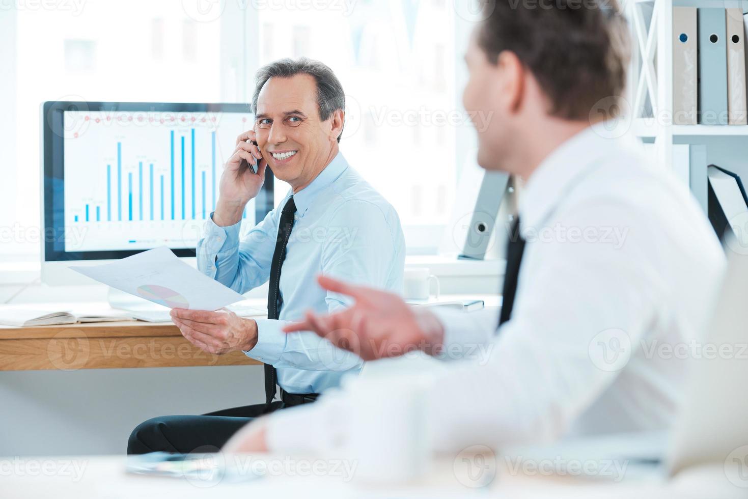 Gut gemacht Zwei fröhliche Geschäftsleute in Abendgarderobe diskutieren etwas und lächeln, während sie im Büro sitzen foto