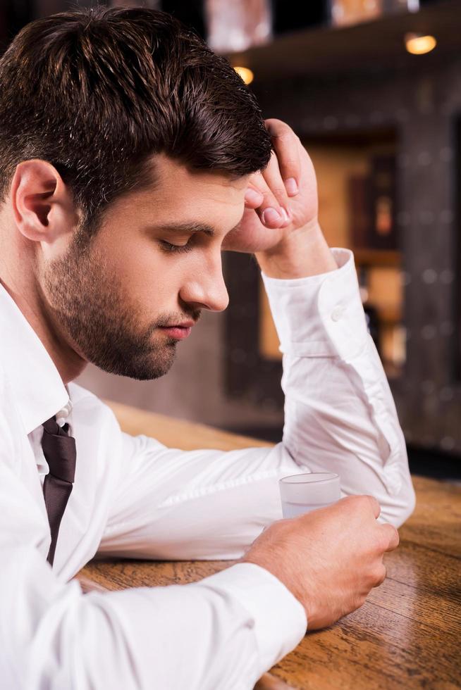 Es war der schlimmste Tag aller Zeiten. Depressiver junger Mann in Hemd und Krawatte, der sich an die Bartheke lehnt und in das Schnapsglas schaut foto