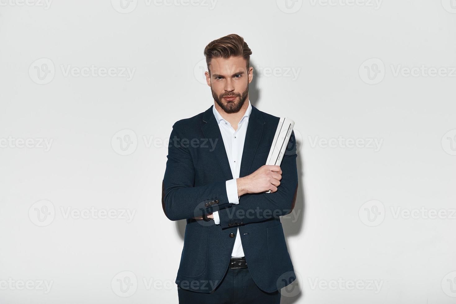 sieht einfach perfekt aus. hübscher junger Mann im vollen Anzug, der die Arme verschränkt hält und in die Kamera schaut, während er vor grauem Hintergrund steht foto