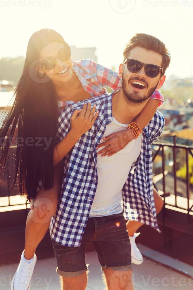 Liebe im Sonnenlicht. Fröhlicher junger Mann, der seine Freundin auf den Schultern trägt, während er gemeinsam Spaß auf dem Dach hat foto