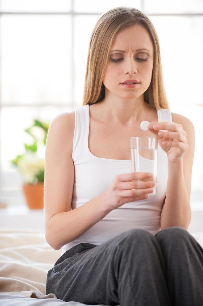 Frau, die Aspirin nimmt. junge Frau hält Pille auf Glas mit Wasser und drückt Negativität aus, während sie im Bett in ihrer Wohnung sitzt foto