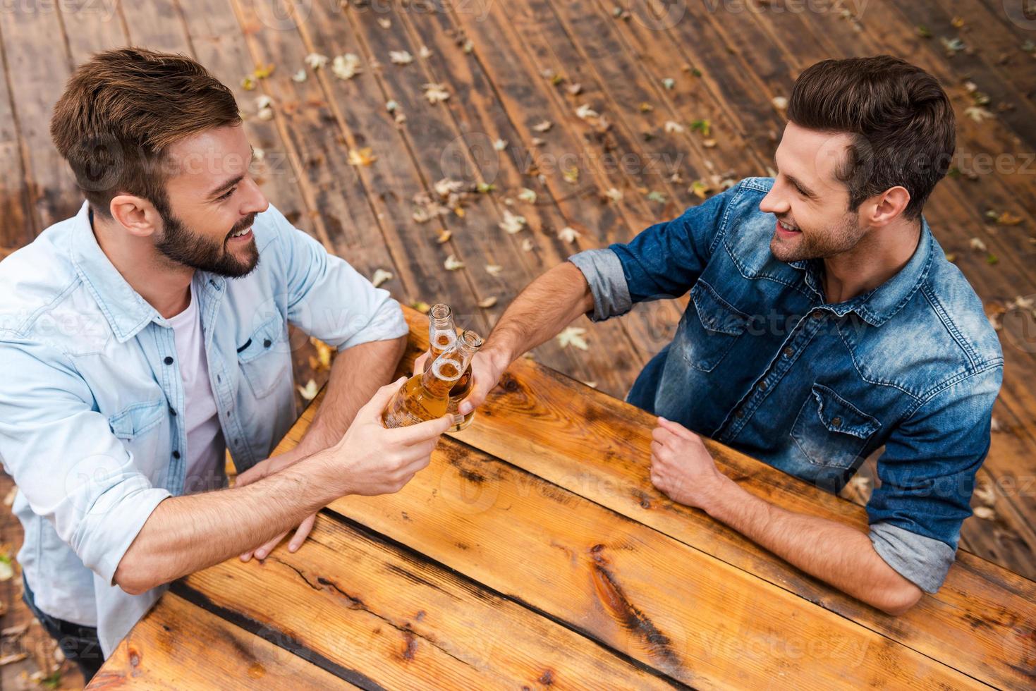 festliche zeit mit der besten freundin. Draufsicht auf zwei fröhliche junge Männer, die mit Biergläsern anstoßen und lächeln, während sie im Freien stehen foto