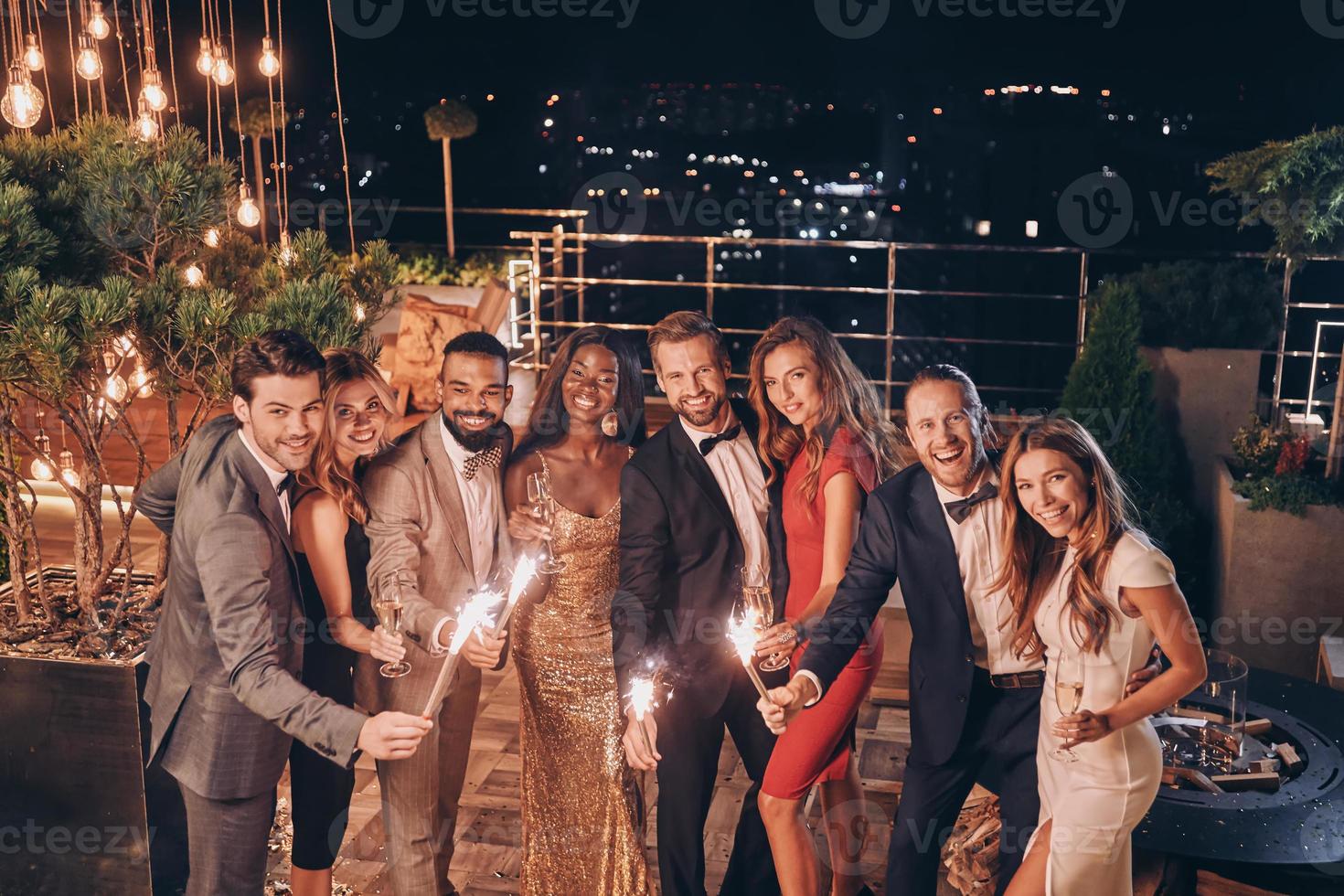 Gruppe schöner Menschen in formeller Kleidung, die Wunderkerzen halten und lächeln, während sie Zeit auf einer Party im Freien verbringen foto