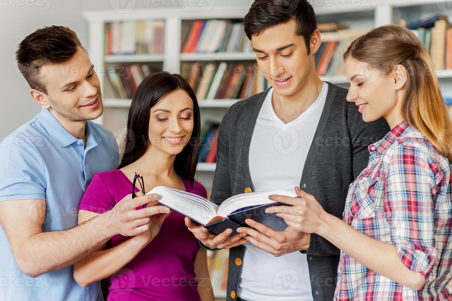 bereiten sich auf ihre Abschlussprüfungen vor. Vier fröhliche Studenten, die zusammen ein Buch lesen, während sie in einer Bibliothek gegen ein Bücherregal stehen foto