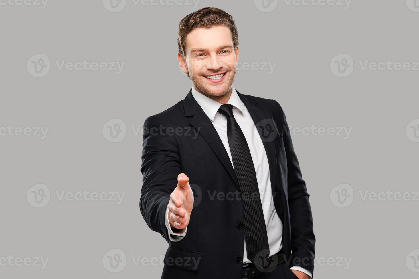 Herzlichen Glückwunsch Porträt eines fröhlichen jungen Mannes in Abendkleidung, der die Hand zum Schütteln ausstreckt, während er vor grauem Hintergrund steht foto