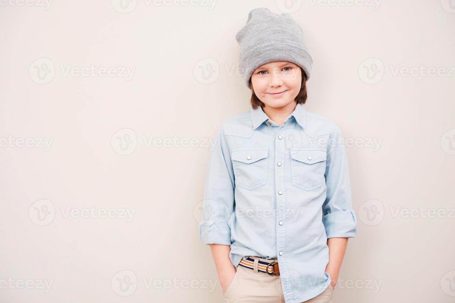 so jung und so stilvoll hübscher kleiner junge mit hut, der hände in den taschen hält und in die kamera schaut, während er vor grauem hintergrund steht foto