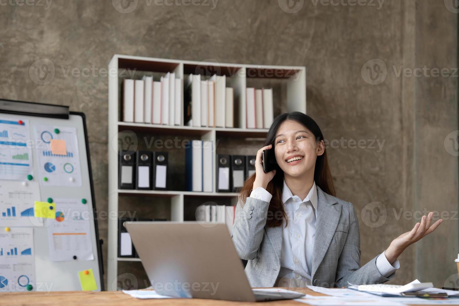 lächelnde schöne asiatische geschäftsfrau, die diagramm und diagramm analysiert, die änderungen auf dem markt zeigen und smartphone im büro halten. foto