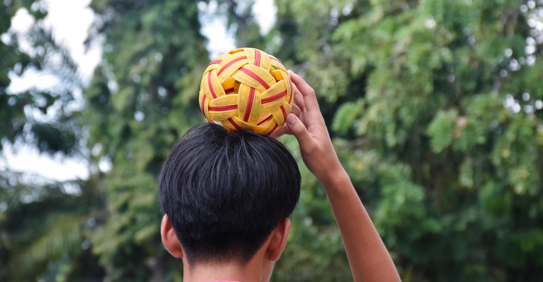 Junger südostasiatischer männlicher Sepak-Takraw-Spieler, der mit seiner rechten Hand den Ball auf dem Kopf hält, Sepak-Takraw-Spiel im Freien nach der Schule, weicher und selektiver Fokus auf den Ball. foto