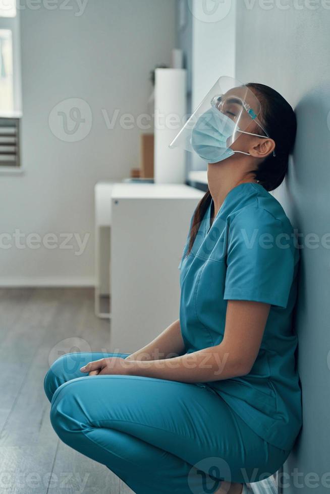 Traurige junge Krankenschwester in Schutzkleidung, die die Augen geschlossen hält, während sie im Krankenhaus arbeitet foto