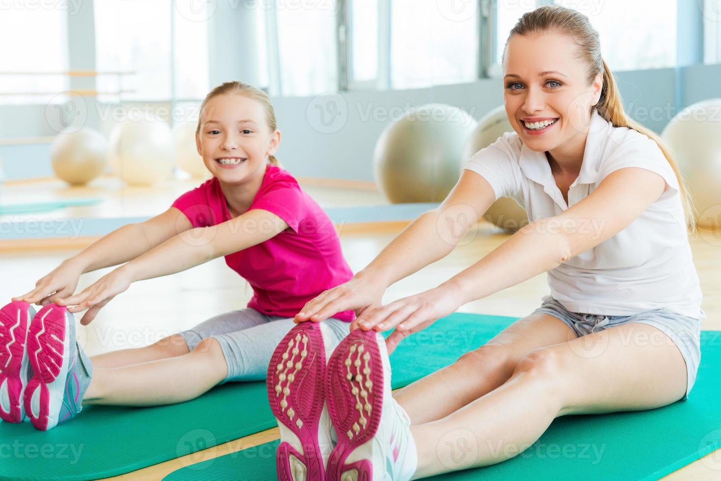 sportliche Mutter und Tochter. fröhliche mutter und tochter, die dehnübungen machen und lächeln, während sie auf übungsmatten im sportverein sitzen foto