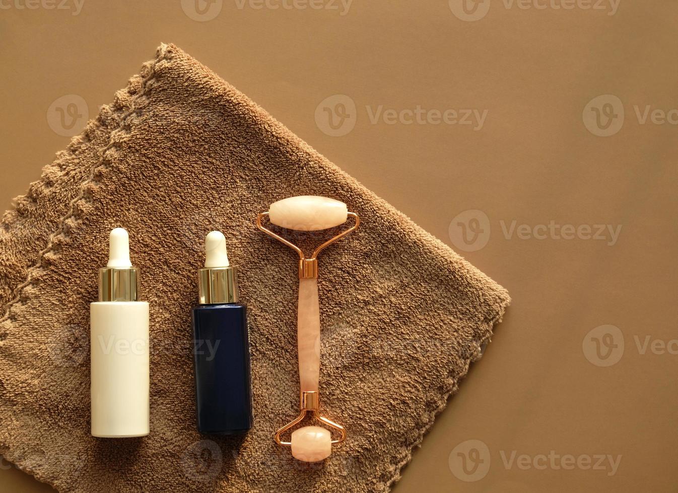 set für die gesichtspflege zu hause auf braunem handtuch. Gesichts-Rosenquarz-Massagerolle oder Gua Sha und Serum in einer weißen Glasflasche und Pipette für die Gesichtspflege. kosmetisches Lifting-Kit, Anti-Aging. foto