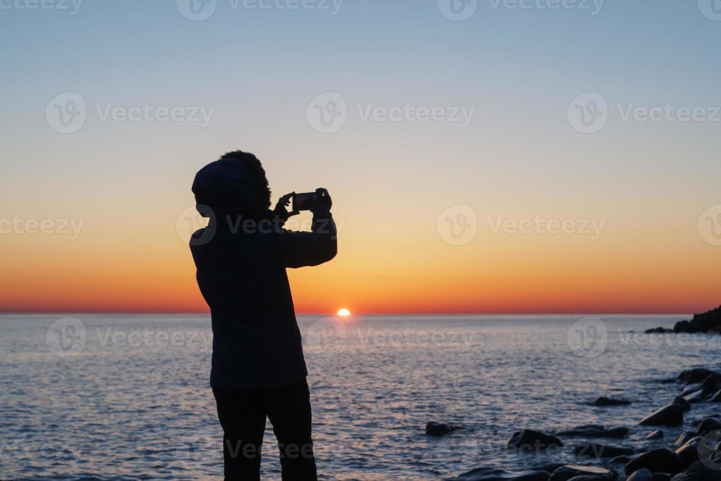 Silhouetten eines Mädchens, das auf einem Smartphone den Sonnenuntergang auf dem Meer fotografiert. foto