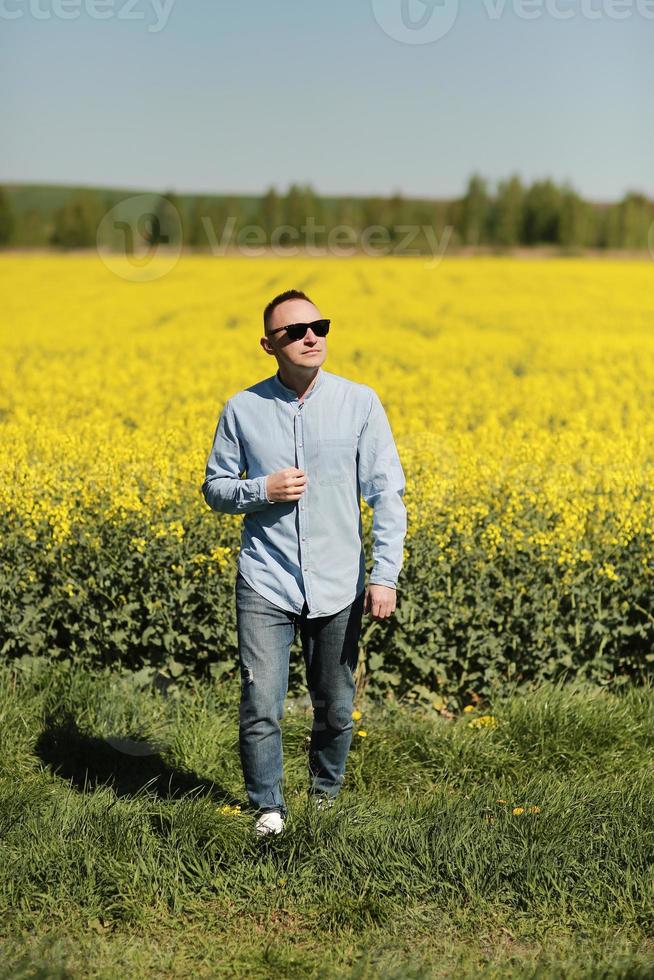 Ein junger Mann mit Sonnenbrille geht spazieren und hat Spaß durch ein sonniges gelbes Rapsfeld, das Konzept von Reisen und Freiheit. sommerferien hintergrund foto