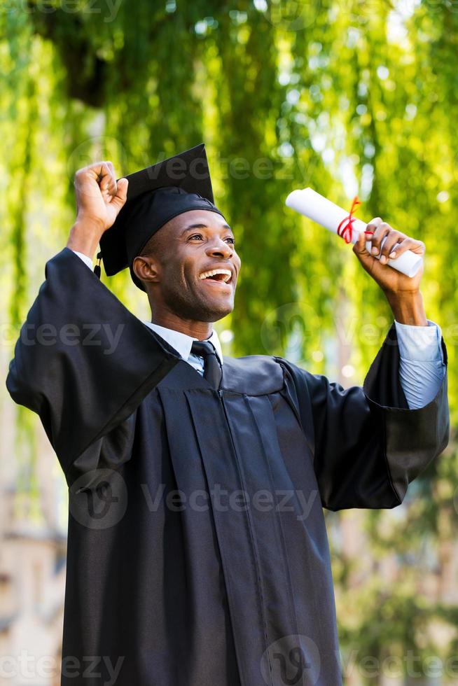 Endlich graduierte glücklicher junger afrikanischer Mann in Abschlusskleidern mit Diplom und erhobenen Armen foto