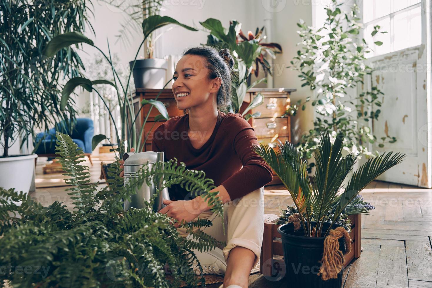 glückliche junge Frau, die Zimmerpflanzen wässert, während sie zu Hause auf dem Boden sitzt foto