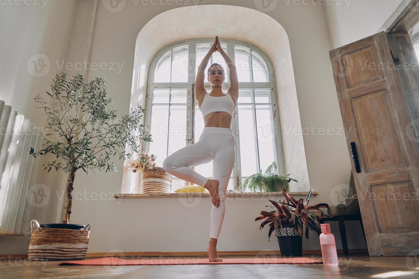 schöne junge frau, die konzentriert aussieht, während sie zu hause yoga vor dem fenster praktiziert foto
