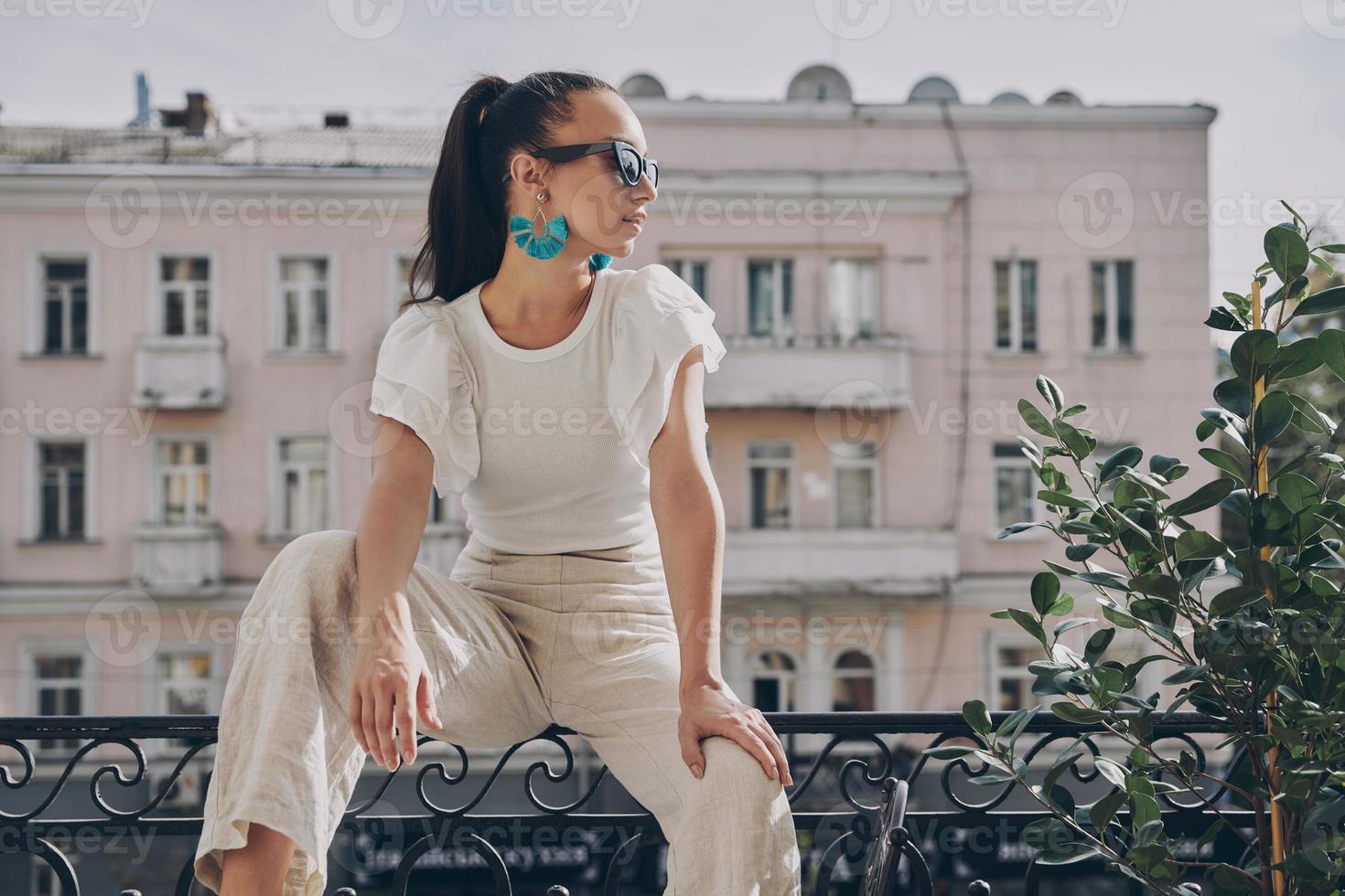 Modische junge Frau mit Brille, die wegschaut, während sie auf dem Balkon sitzt foto