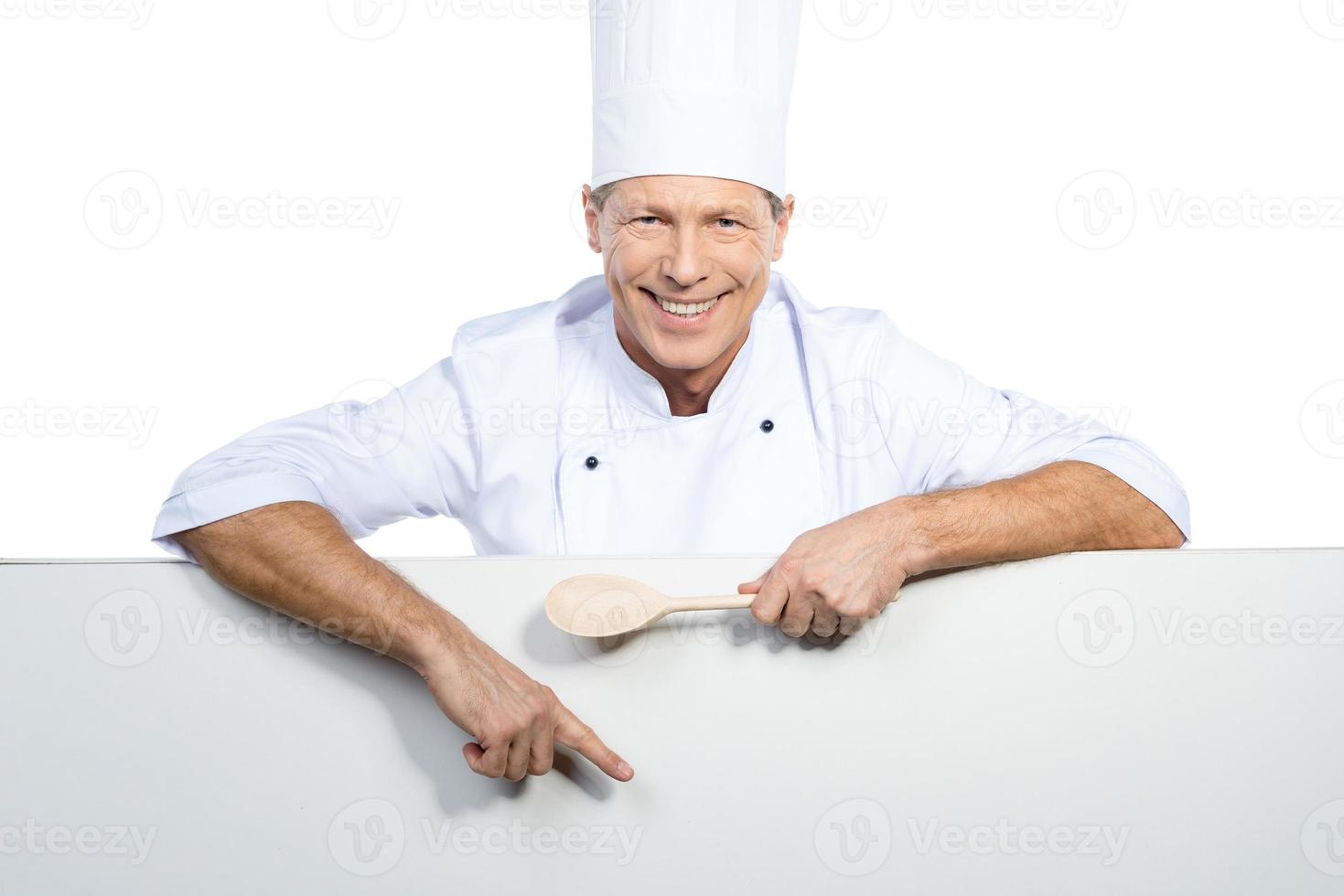 koch zeigt kopierraum. Selbstbewusster, reifer Koch in weißer Uniform, der einen Holzlöffel hält und Kopienraum zeigt, während er vor weißem Hintergrund steht foto