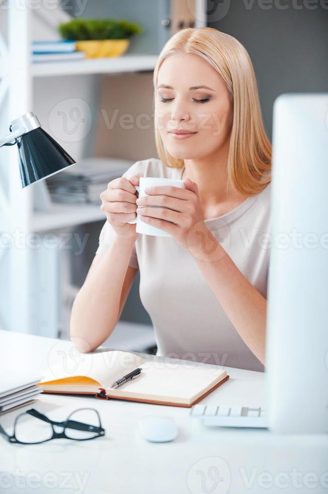 was für ein schöner tag schöne junge frau, die tasse mit heißem getränk hält und die augen geschlossen hält, während sie an ihrem arbeitsplatz im büro sitzt foto