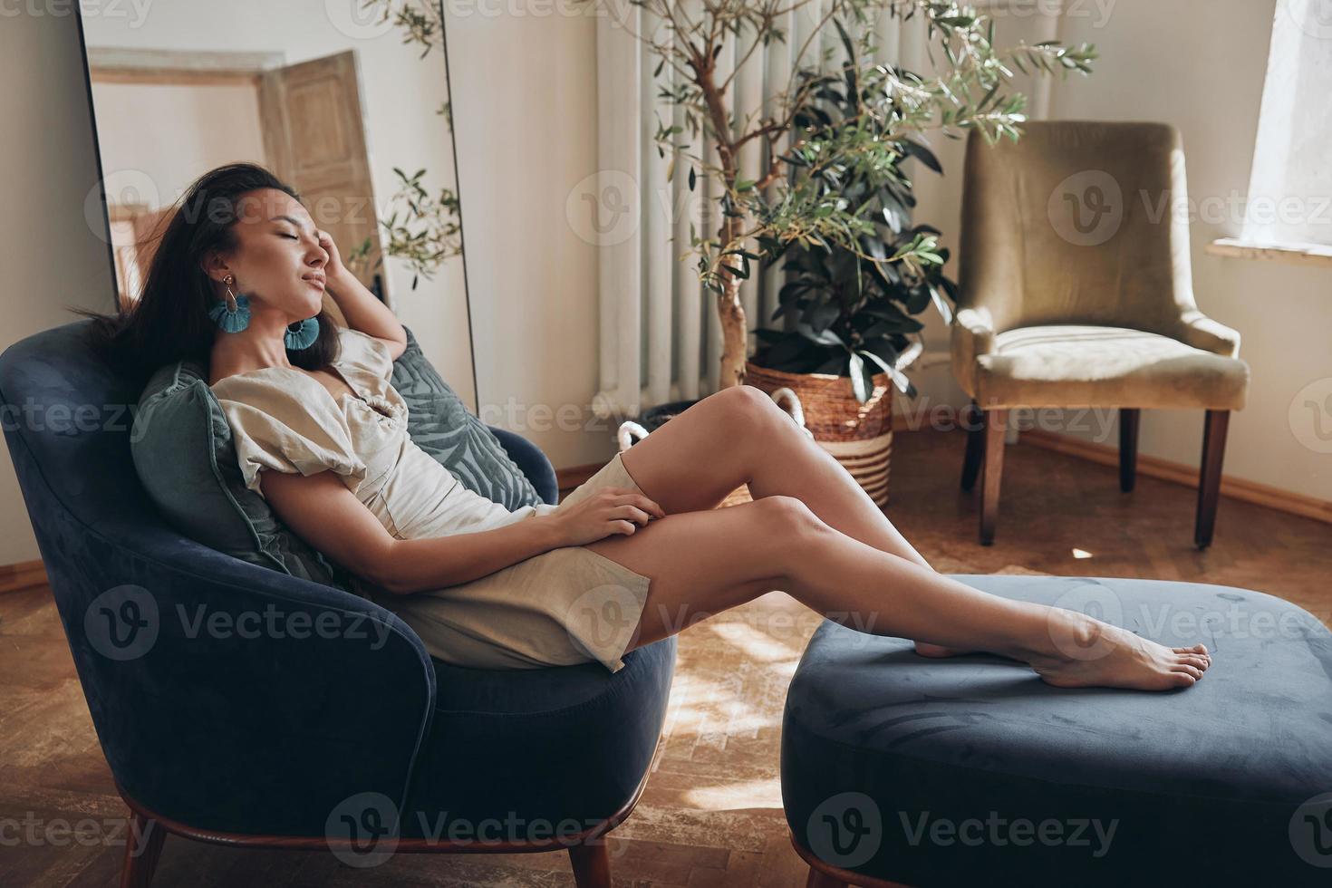 Schöne junge Frau, die die Augen geschlossen hält, während sie sich zu Hause in einem bequemen Stuhl entspannt foto