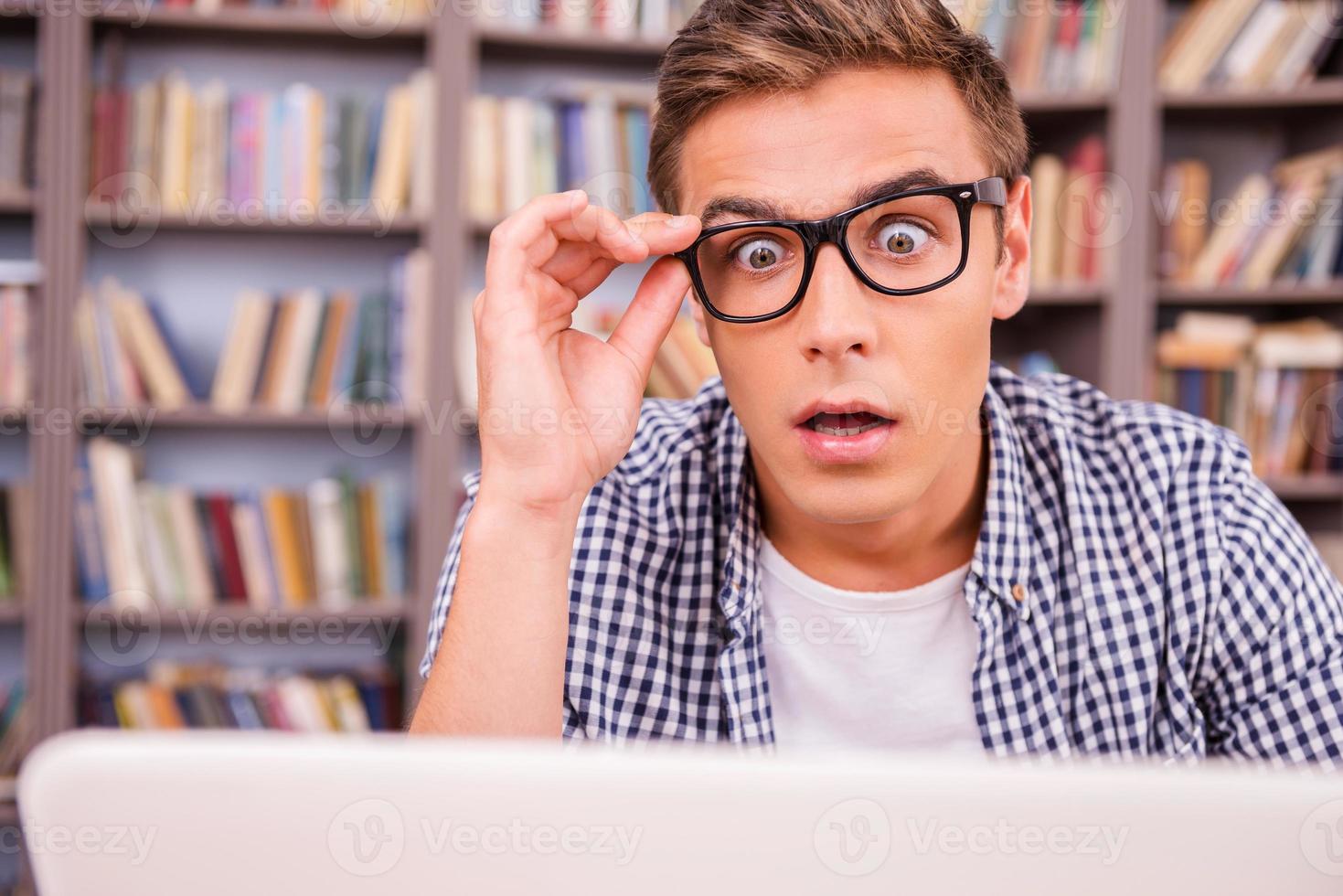 Es ist unglaublich überrascht, wie ein junger Mann auf einen Laptop schaut und den Mund offen hält, während er gegen ein Bücherregal sitzt foto