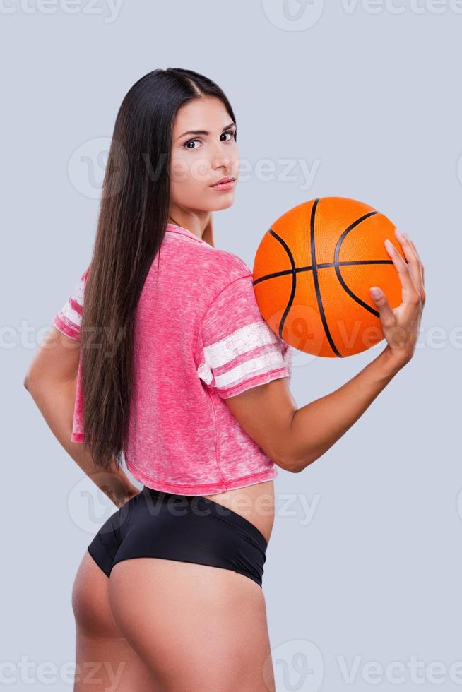 ich liebe Basketball schöne junge Cheerleaderin, die Basketballball hält und über die Schulter schaut, während sie vor grauem Hintergrund steht foto