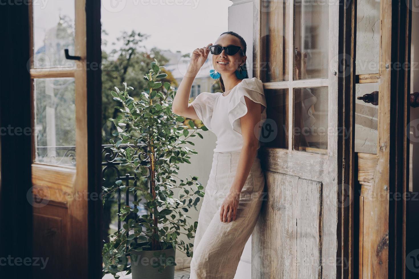 Modische junge Frau, die ihre Brille anpasst und lächelt, während sie auf dem Balkon steht foto