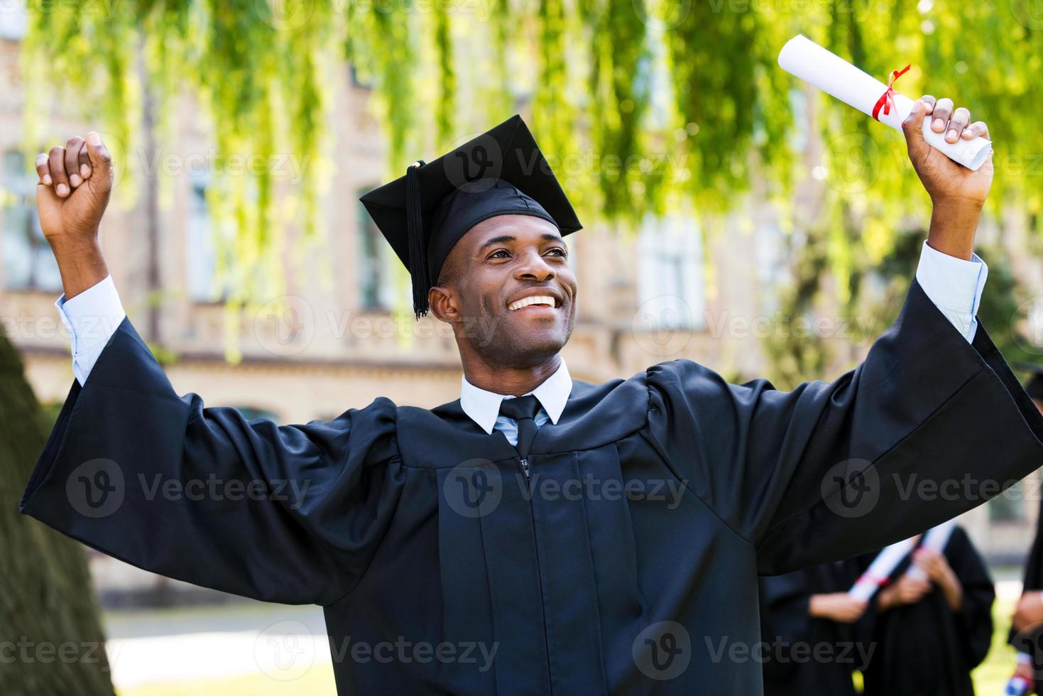 Ich habe endlich meinen Abschluss gemacht, ein glücklicher junger afrikanischer Mann in Abschlusskleidern, der sein Diplom in der Hand hält und die Arme hochhebt, während seine Freunde im Hintergrund stehen foto