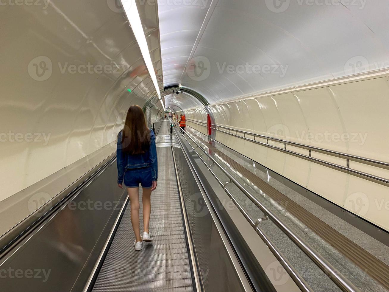 Ein schönes Mädchen geht den Travolator im U-Bahn-Tunnel entlang, um zu einer anderen U-Bahn-Station zu gelangen. Ansicht von hinten foto