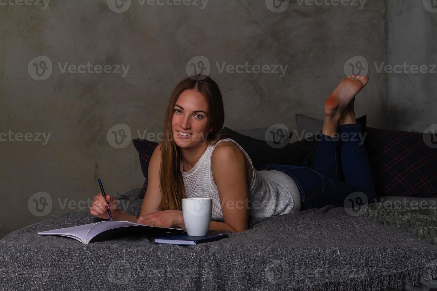 lächelnde Frau mit einem Notizbuch und einer weißen Tasse, die auf dem Bauch im Bett liegt foto