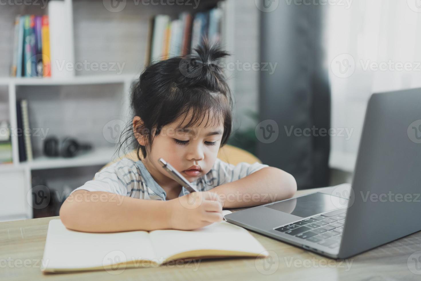 asiatisches baby, das ein blau gestreiftes hemd trägt, benutzt laptop und schreibt notizen in notizbuch, um online auf holztischschreibtisch im wohnzimmer zu hause zu lernen. bildung lernen online von zu hause aus. foto