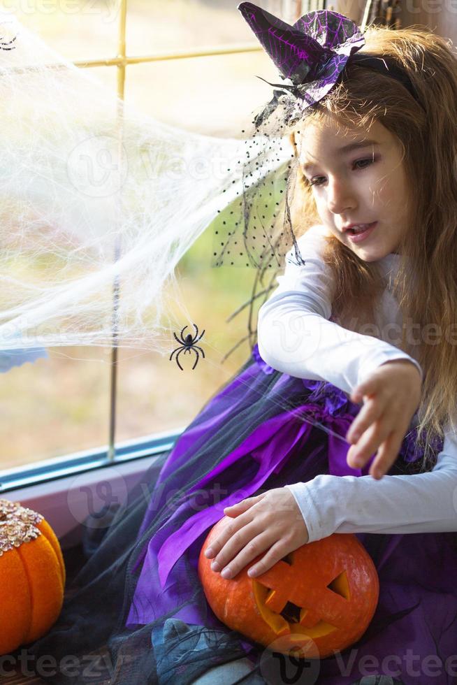 ein mädchen in einem hexenkostüm auf der fensterbank am fenster spielt mit einem spinnennetz und einer spinne und schmückt das haus für den halloween-urlaub. schreckliche Landschaften, Angst und Schrecken, Kürbisse foto