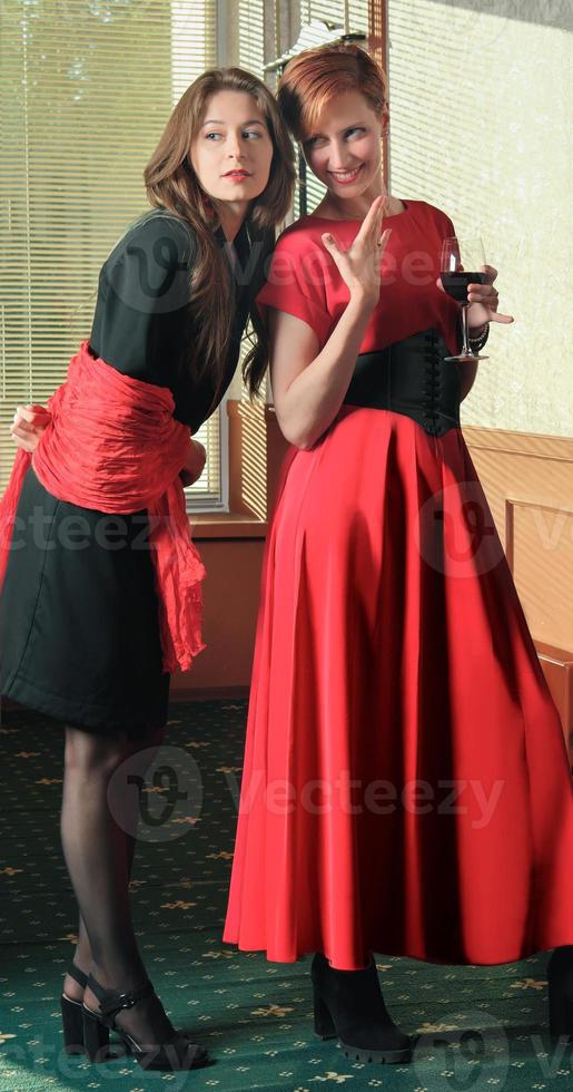 Ganzkörperporträt eleganter junger Frauen mit Weingläsern auf der Feierparty. schwarzes und rotes Outfit. foto