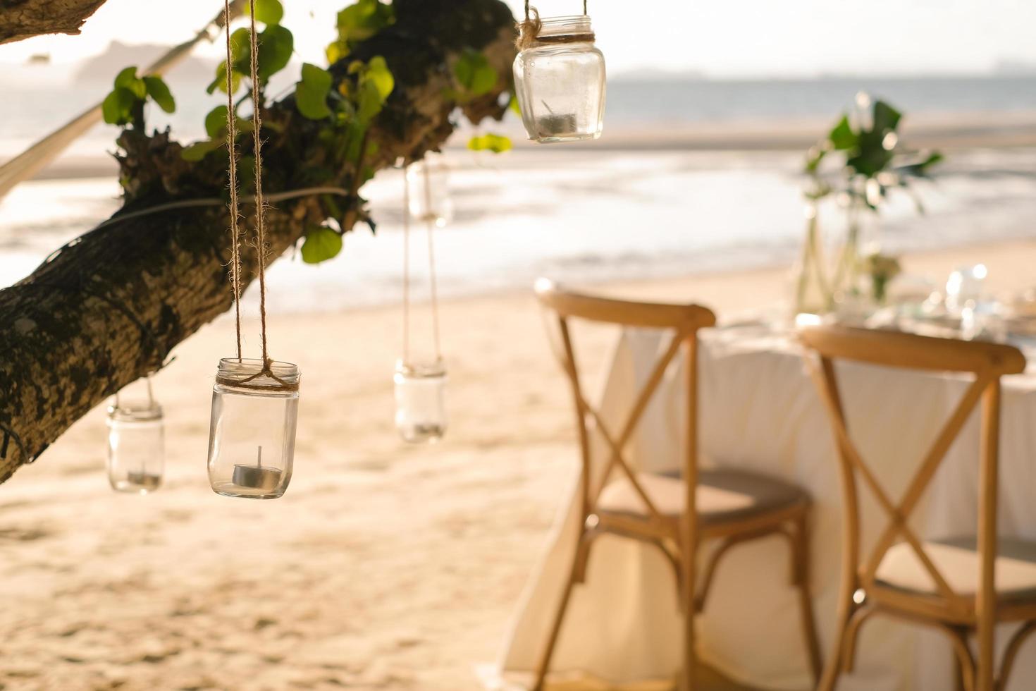 schließen sie die glaskerze, die am baum hängt, mit einem langen tisch hochzeitsessen am strand von thailand am abend. Hochzeitsfeier-Konzept. Dekoration Restaurant im Freien am Strand. foto