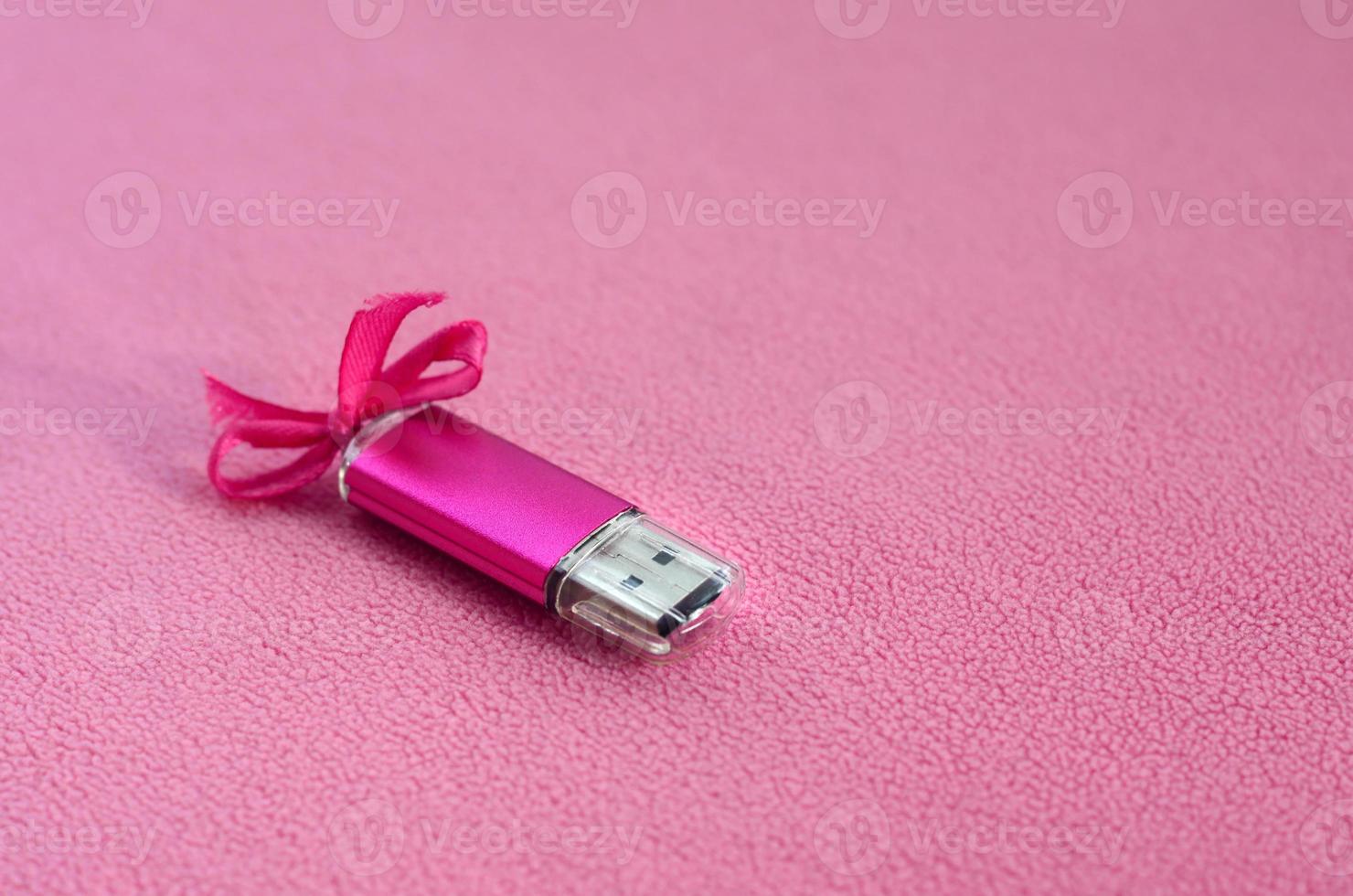 Eine leuchtend rosa USB-Flash-Speicherkarte mit einer rosa Schleife liegt auf einer Decke aus weichem und pelzigem hellrosa Fleece-Stoff. klassisches weibliches Geschenkdesign für eine Speicherkarte foto