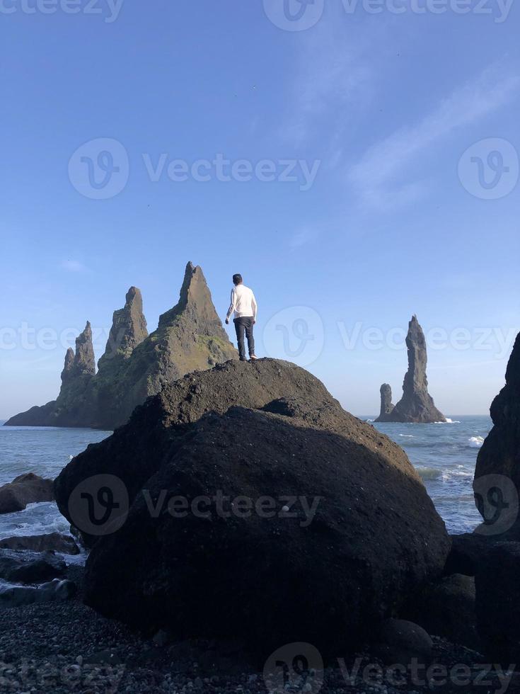 Mann steht auf einem Felsen am schwarzen Strand von Reynisfjara, Island, mit Wind im Haar foto
